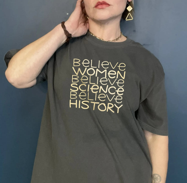 A woman wearing a gray t-shirt with the text "Believe Women Believe Science Believe History" printed on it.
