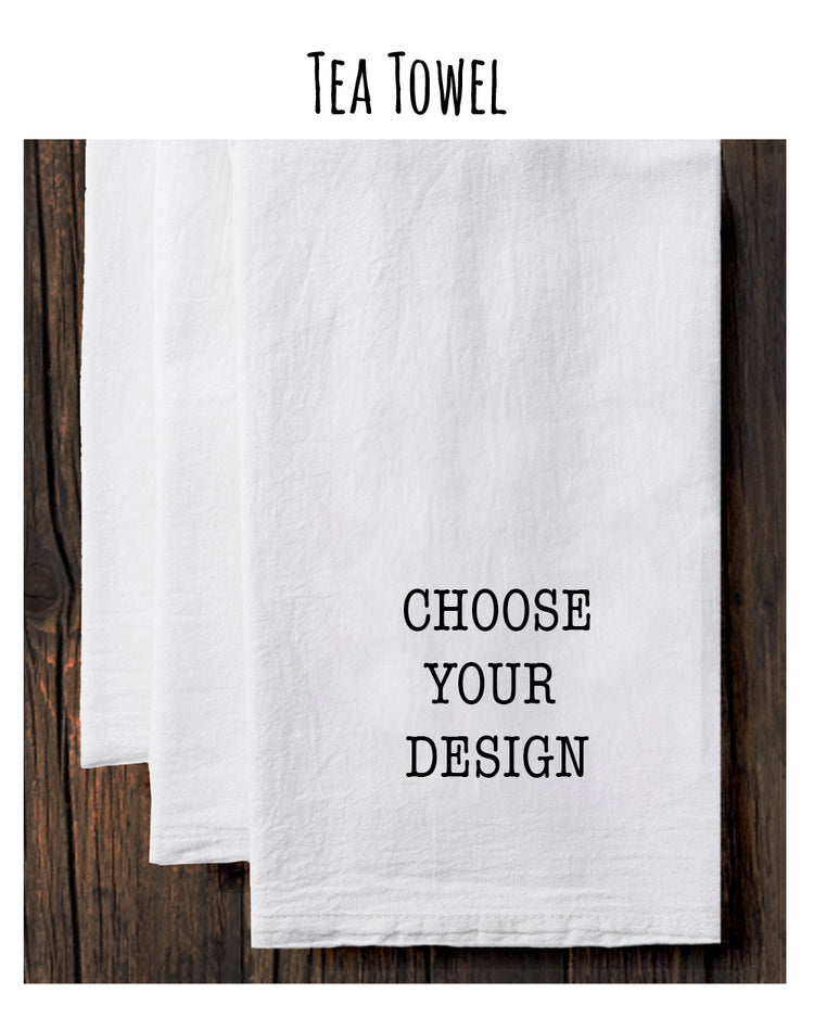 A white tea towel with the text "CHOOSE YOUR DESIGN" printed on it, placed on a wooden surface.