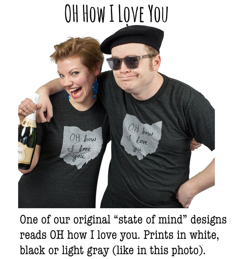 A man and a woman wearing matching black t-shirts with handwritten text, posing together and smiling.