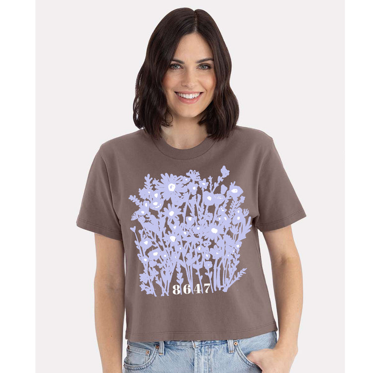 A woman wearing a brown t-shirt with a graphic design of purple flowers and the number "8647" printed on it.