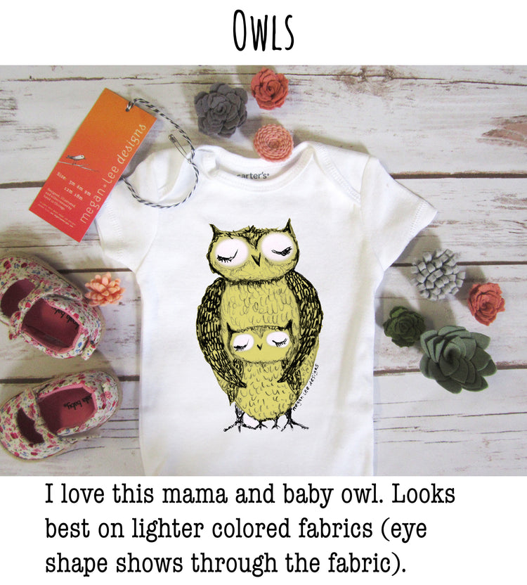 a white baby onesie with a colorful owl design, placed on a wooden surface alongside other items such as a pair of baby shoes, flowers, and a tag.