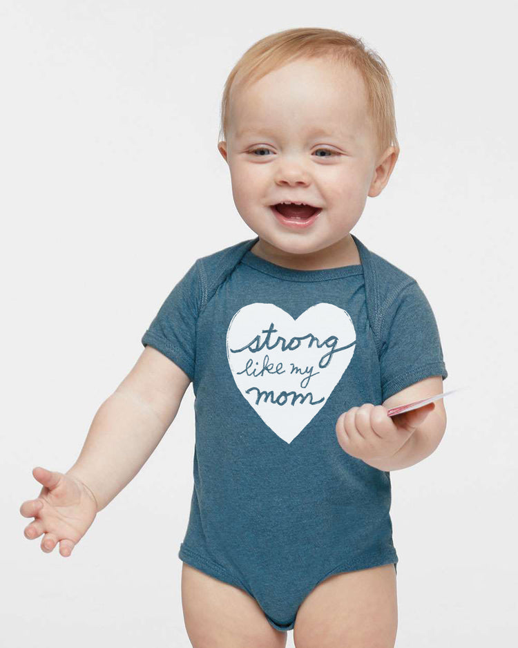 A smiling baby boy wearing a blue onesie with a heart and the text "Strong like my mom" printed on it.