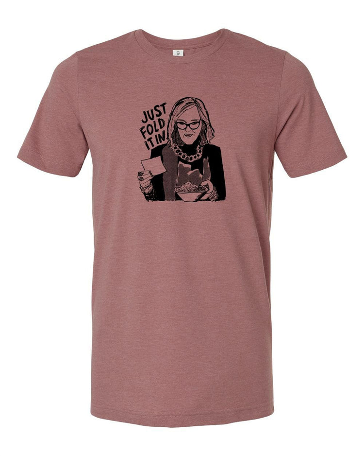 A graphic t-shirt design featuring a woman with glasses and a serious expression, holding a book and a cup of coffee. The text above the image says "JUST FOLD IT".