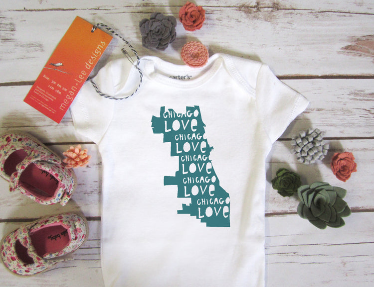 A white baby onesie with a graphic of Chicago, surrounded by various baby items such as shoes, flowers, and other decorative objects.