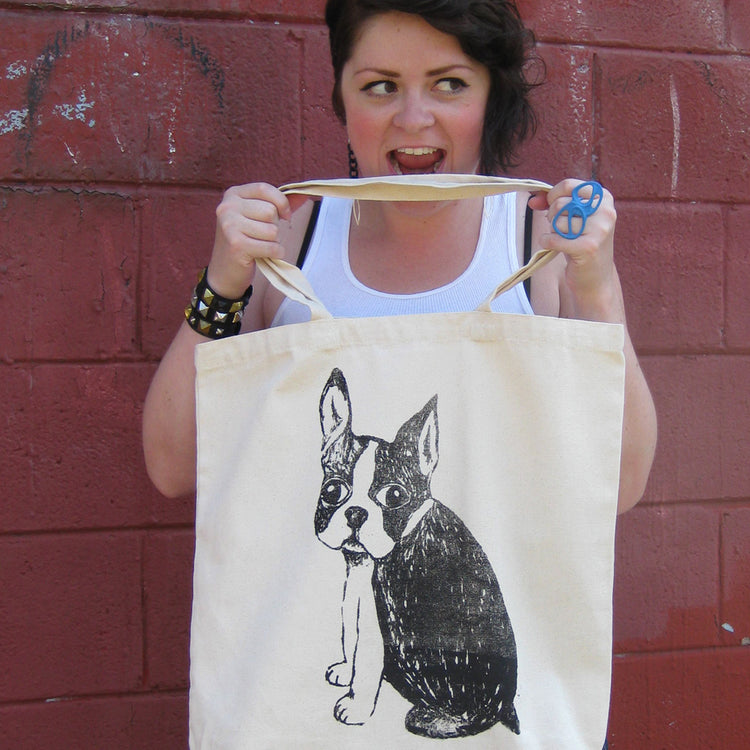 A woman is holding a white tote bag with a black and white dog illustration on it, and she is making a funny or surprised face.