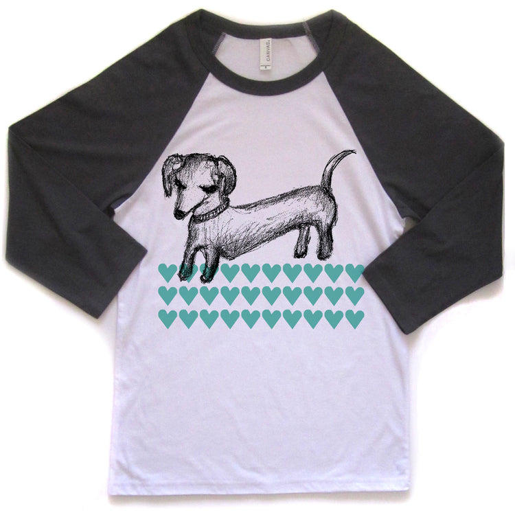 A white and black baseball-style t-shirt with a graphic design of a dachshund dog and a pattern of green hearts.
