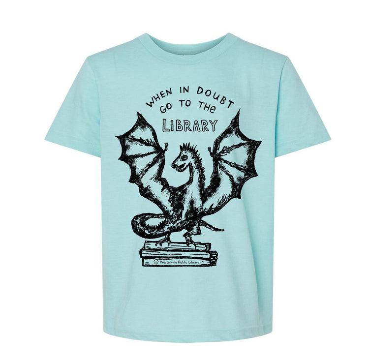 A light blue t-shirt with a graphic design depicting a dragon perched on a stack of books, accompanied by the text "WHEN IN DOUBT GO TO THE LIBRARY" above the dragon.