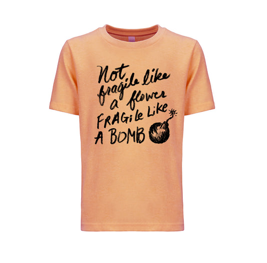 The image is a peach-colored t-shirt with a black text design printed on it. The text reads "Not, fragile like a flower, fragile like a bomb", accompanied by a graphic of a bomb.