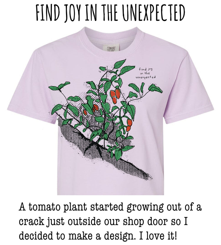 A pink t-shirt with a graphic of a tomato plant growing out of a crack on the front, accompanied by the text "Find joy in the unexpected" and a caption describing how a tomato plant started growing out of a crack outside their shop door, leading to the creation of a design.