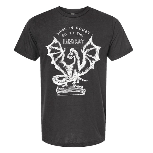 A black t-shirt with a white graphic design depicting a dragon with wings spread wide, standing on top of a stack of books. The text above the dragon reads "WHEN IN DOUBT, GO TO THE LIBRARY".