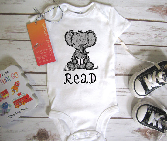 A baby onesie with the word "Read" printed on it, along with a pair of black and white sneakers, a children's book, and a tag.