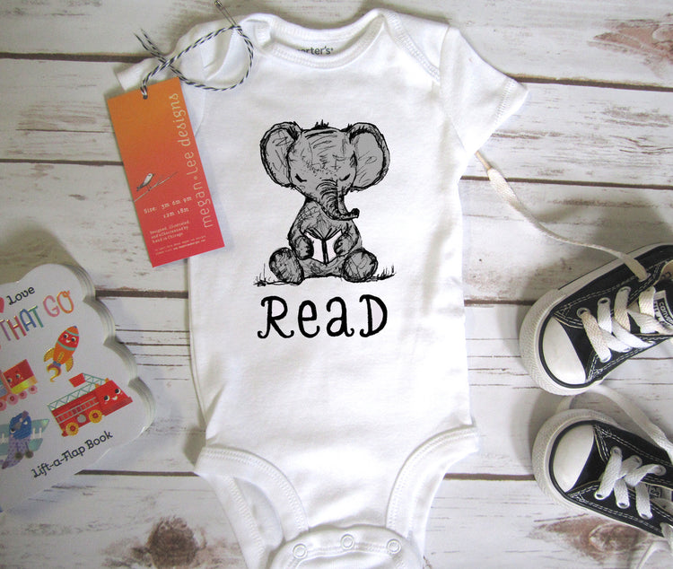 A baby onesie with the word "Read" printed on it, along with a pair of black and white sneakers, a children's book, and a tag.