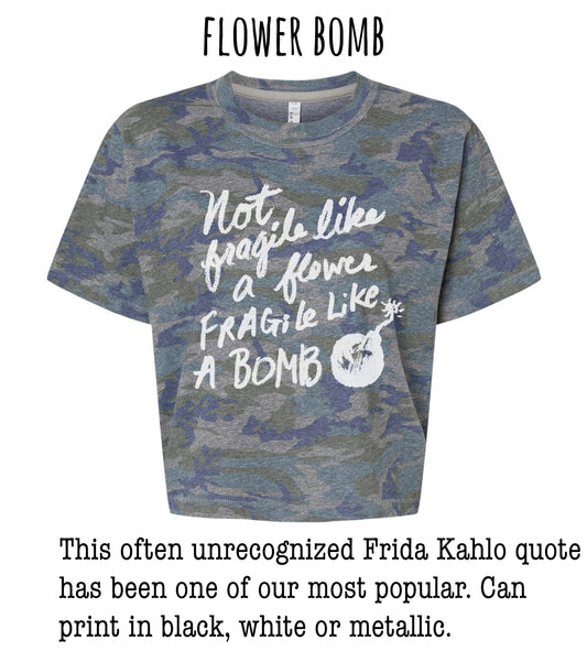 a gray camouflage t-shirt with a white text quote that reads "Not like a flower, fragile like a bomb" and a small white explosion symbol. The quote is often unacknowledged and has been one of our most popular. The t-shirt can be printed in black, white, or metallic colors.
