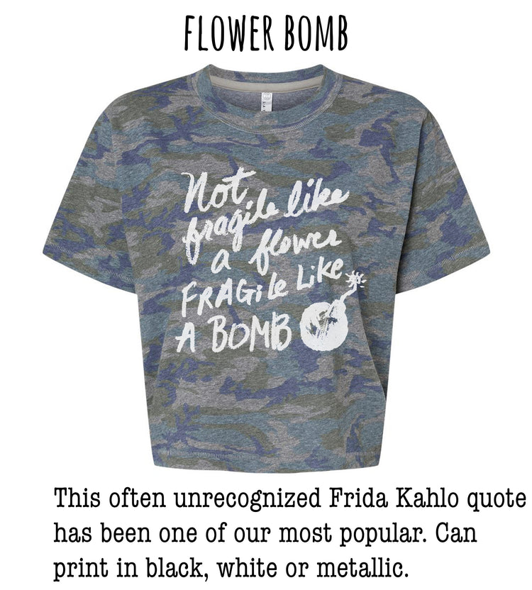 a gray camouflage t-shirt with a white text quote that reads "Not like a flower, fragile like a bomb" and a small white explosion symbol. The quote is often unacknowledged and has been one of our most popular. The t-shirt can be printed in black, white, or metallic colors.