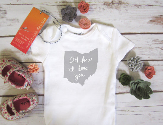 a white baby onesie with the text "OH how I love you" printed on it, along with a pair of floral baby shoes and some decorative flowers on a wooden surface.
