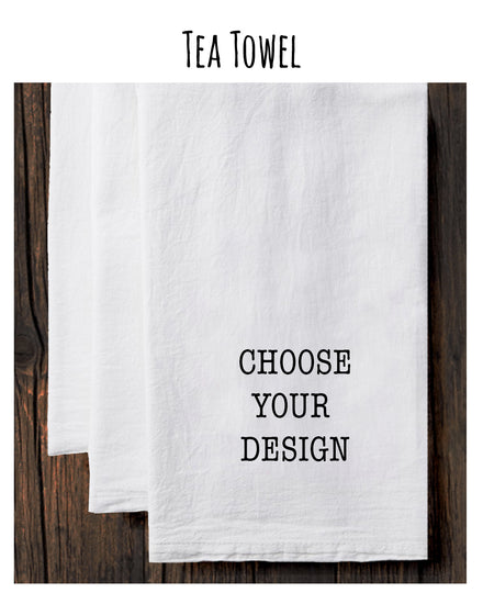 A white tea towel with the text "CHOOSE YOUR DESIGN" printed on it, placed on a wooden surface.