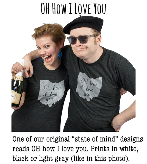 A man and a woman wearing matching black t-shirts with handwritten text, posing together and smiling.