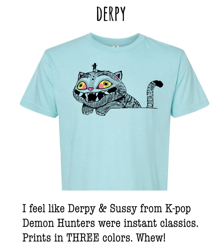 A light blue t-shirt with a cartoon cat graphic and text that says "DERPY I feel like Derpy & Susy from K-pop Demon Hunters were instant classics. Prints in THREE colors. Whew!"