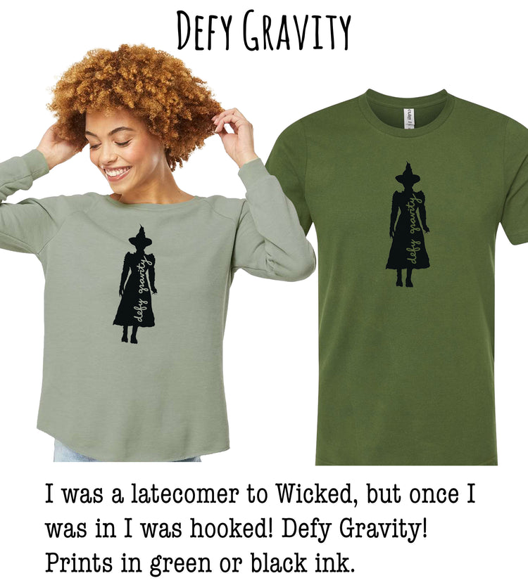 A woman with curly hair is wearing a green t-shirt with a black silhouette of a person wearing a witch's hat. The t-shirt has the text "Defy Gravity" printed on it.