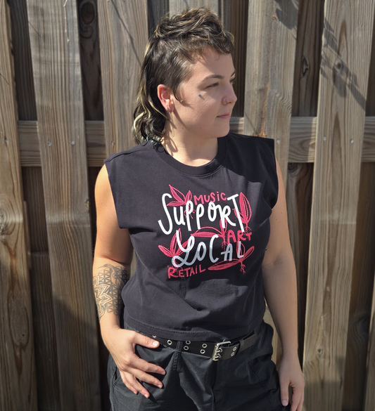 A person wearing a sleeveless black t-shirt with text printed on it, standing in front of a wooden fence.