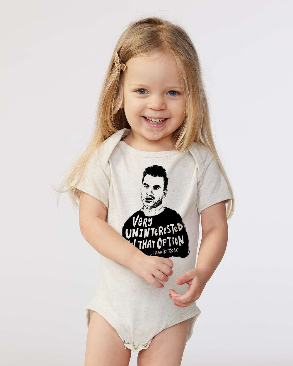 A young girl with blonde hair is smiling and wearing a white onesie with a graphic design and text printed on it.