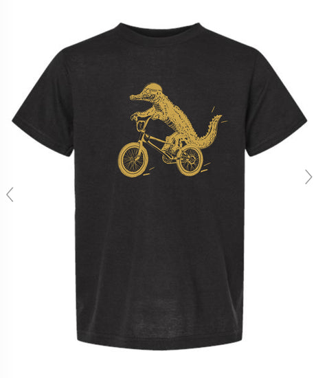 A black t-shirt with a gold graphic design depicting a crocodile riding a bicycle.