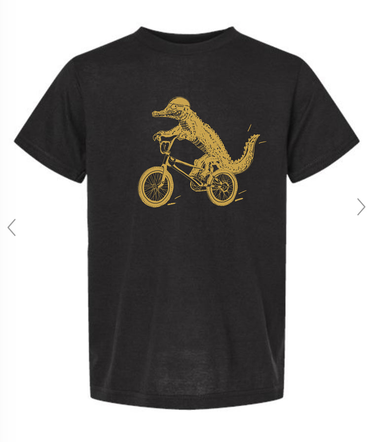 A black t-shirt with a gold graphic design depicting a crocodile riding a bicycle.