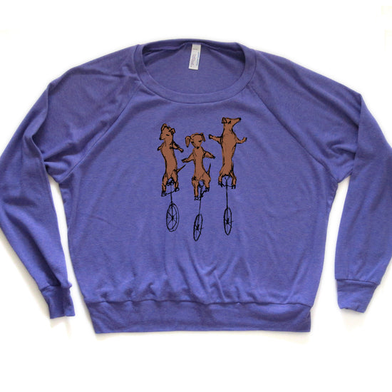 A purple long-sleeved shirt with a graphic design depicting three brown dogs riding on unicycle wheels.