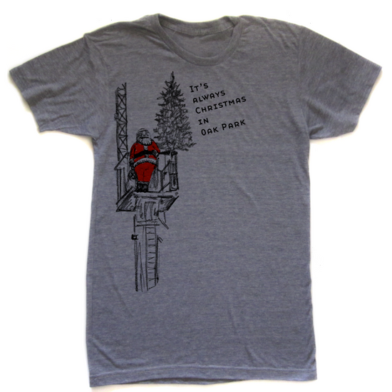A gray t-shirt with a graphic design depicting a person in a red suit standing on a platform, with a Christmas tree in the background. The text on the t-shirt reads "It's always Christmas in Oak Park".