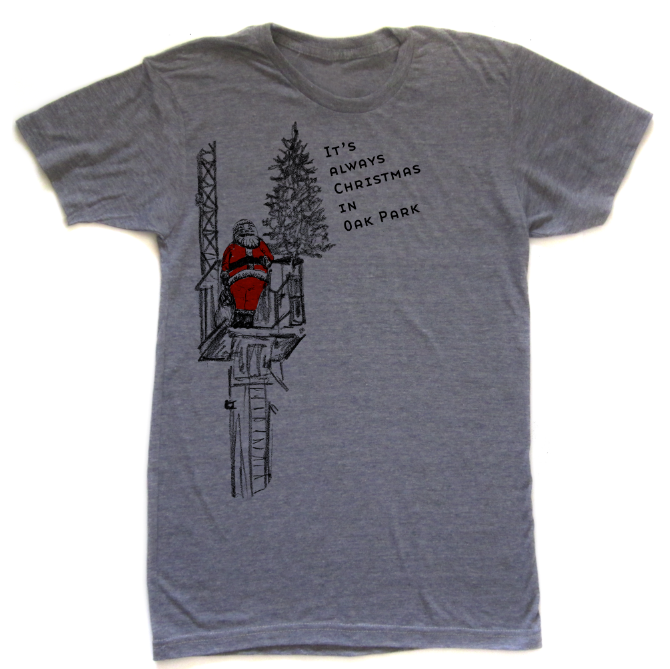 A gray t-shirt with a graphic design depicting a person in a red suit standing on a platform, with a Christmas tree in the background. The text on the t-shirt reads "It's always Christmas in Oak Park".