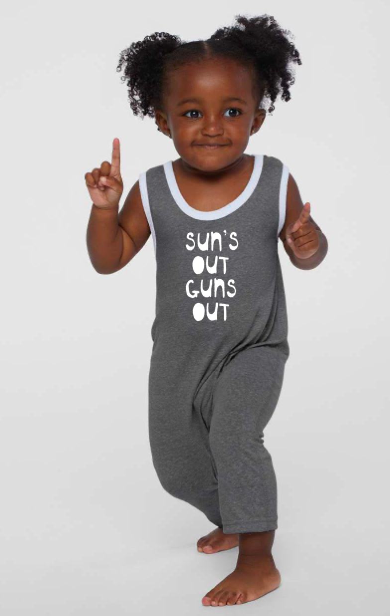 A young girl with curly hair is standing and pointing upwards, wearing a gray jumpsuit with the text "Sun's out guns out" printed on it.