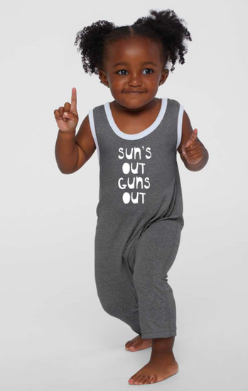A young girl with curly hair is standing and pointing upwards, wearing a gray jumpsuit with the text "Sun's out guns out" printed on it.