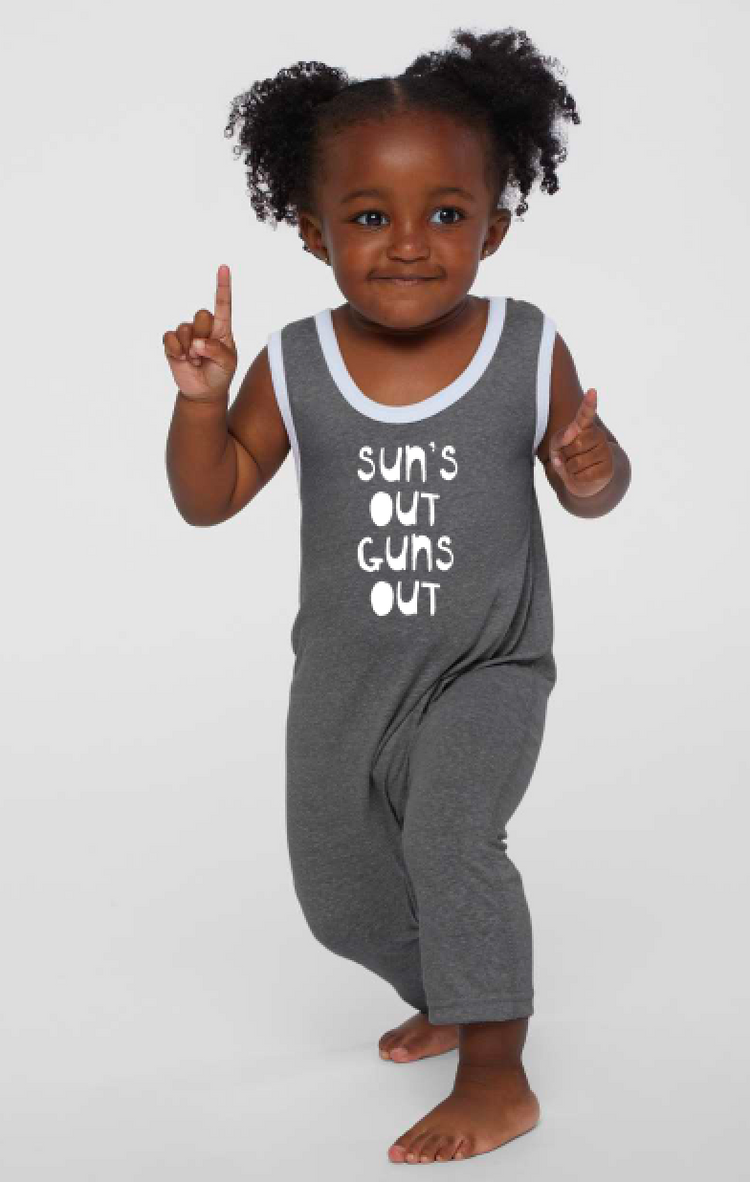 A young girl with curly hair is standing and pointing upwards, wearing a gray jumpsuit with the text "Sun's out guns out" printed on it.