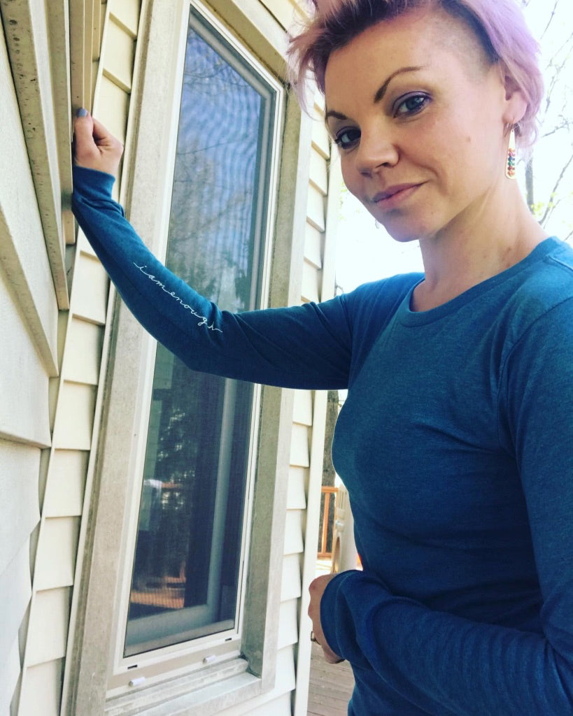 A woman with short hair and a blue shirt stands in front of a window, looking directly at the camera with a serious expression.