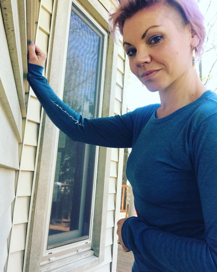 A woman with short hair and a blue shirt stands in front of a window, looking directly at the camera with a serious expression.