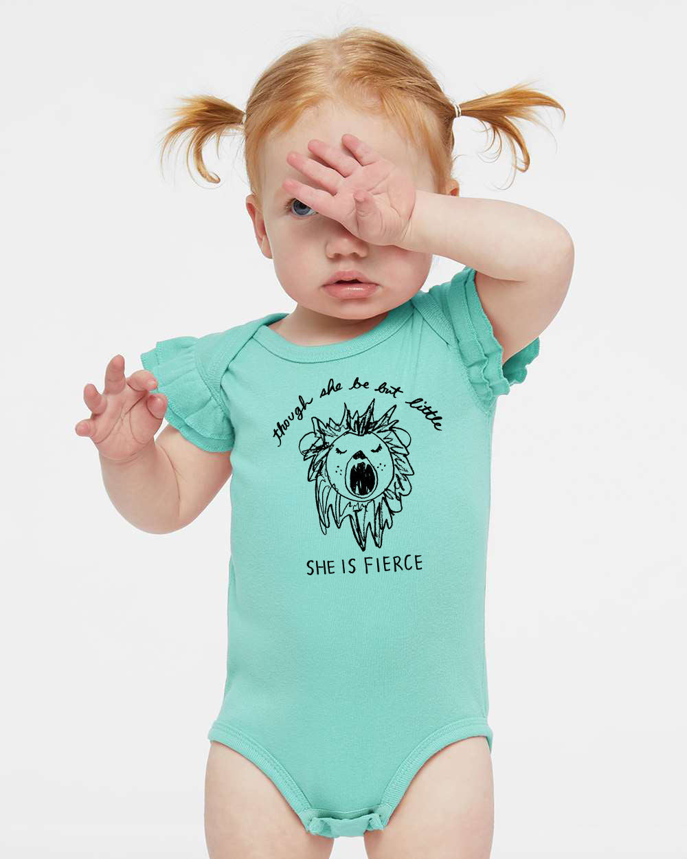 A young child with red hair wearing a light blue onesie with a lion graphic and the text "She is fierce" printed on it.