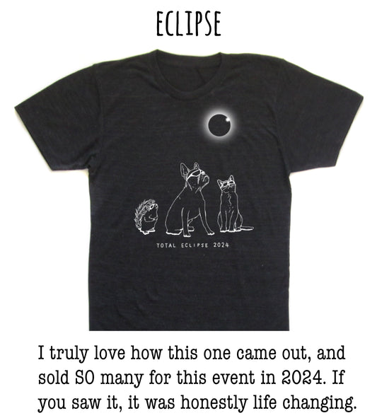 The image is a black t-shirt with a graphic design depicting a total eclipse, featuring a moon and three animals - a dog, a cat, and a hedgehog - against a dark background.