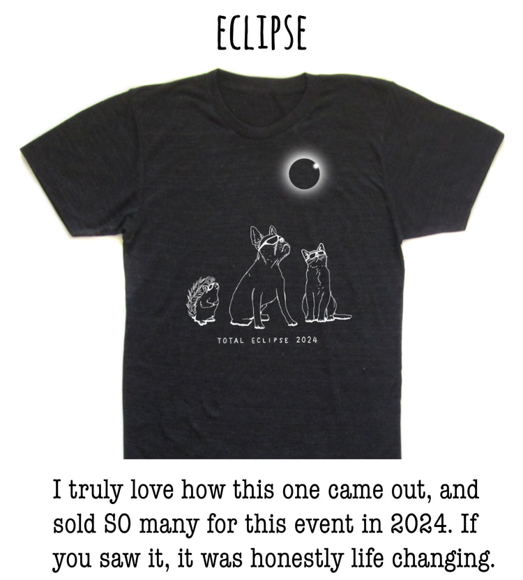 The image is a black t-shirt with a graphic design depicting a total eclipse, featuring a moon and three animals - a dog, a cat, and a hedgehog - against a dark background.