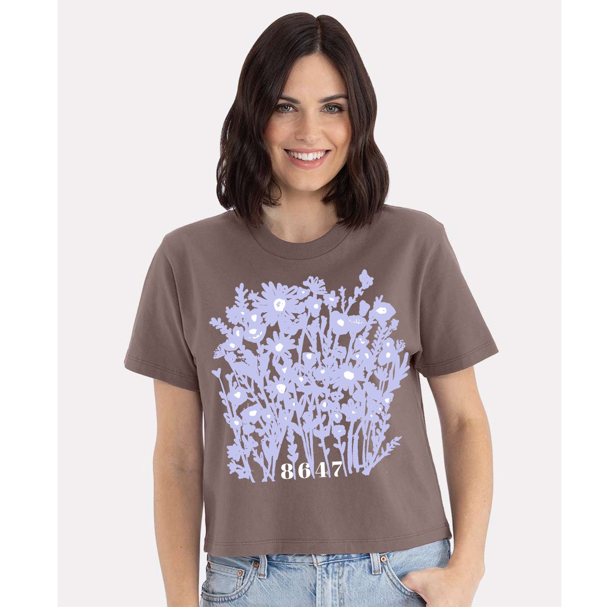 A woman wearing a brown t-shirt with a graphic design of purple flowers and the number "8647" printed on it.
