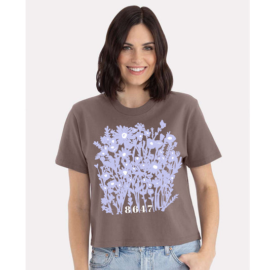 A woman wearing a brown t-shirt with a graphic design of purple flowers and the number "8647" printed on it.