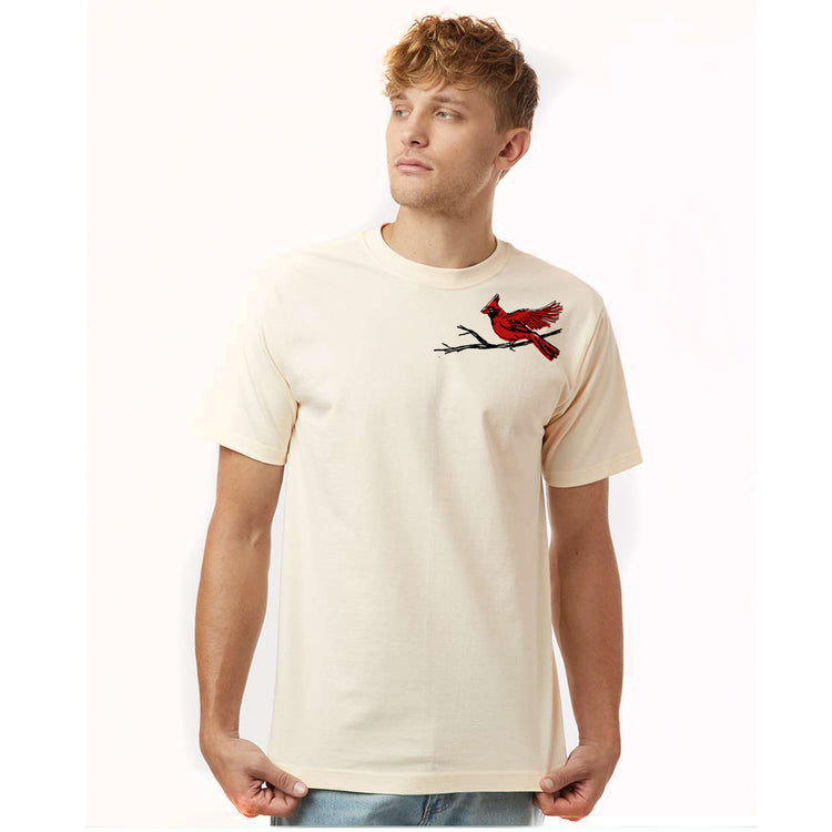 A young man wearing a cream-colored t-shirt with a red bird design on the left side of his chest.