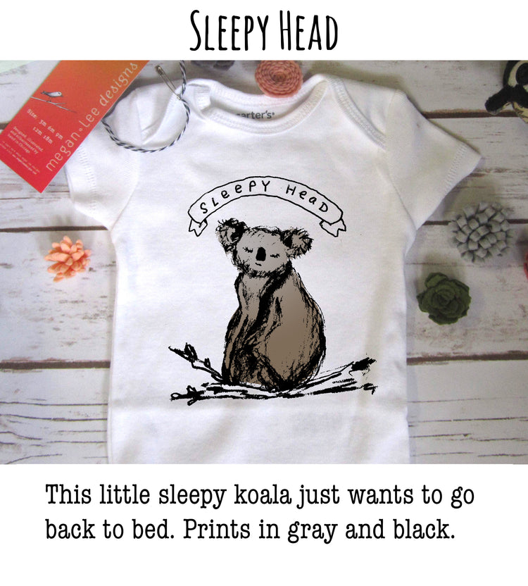 A white baby onesie with a black and white illustration of a koala bear and the text "SLEEPY HEAD" printed on it, along with a small red tag and some decorative items.