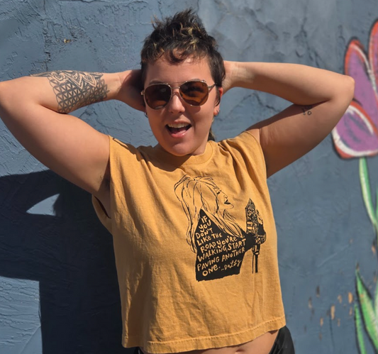 A person wearing a yellow t-shirt with a graphic design and sunglasses stands in front of a graffiti-covered wall.