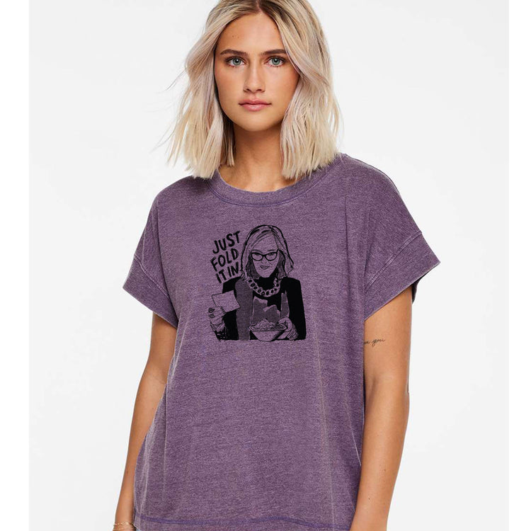 A woman with blonde hair wearing a purple t-shirt with a graphic design of a woman holding a sign that says "Just fold it".