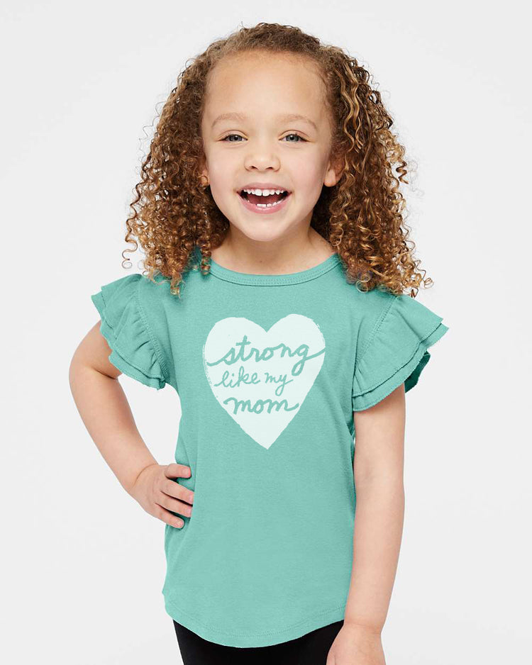 A young girl with curly hair is smiling and wearing a teal-colored t-shirt with a heart design and the text "Strong like my mom" printed on it.