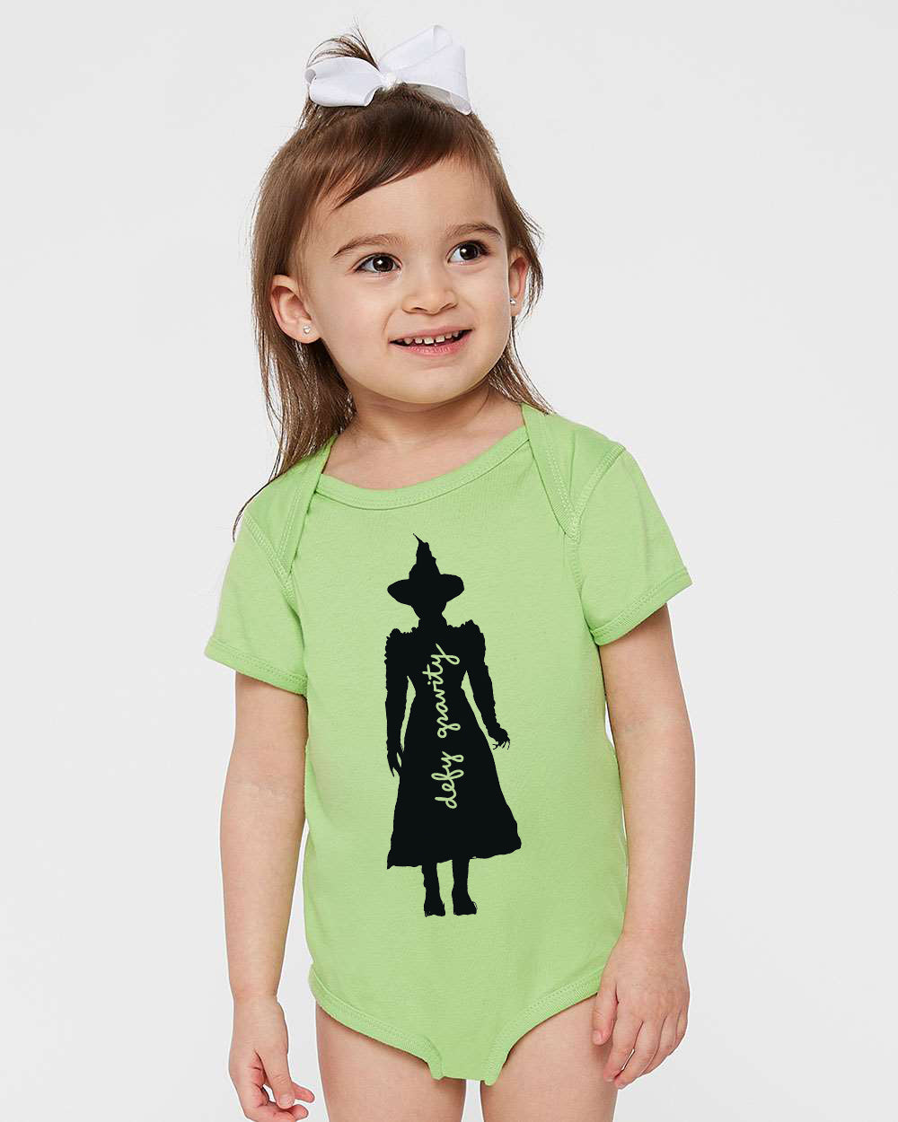 A young girl wearing a green onesie with a silhouette of a witch on it.