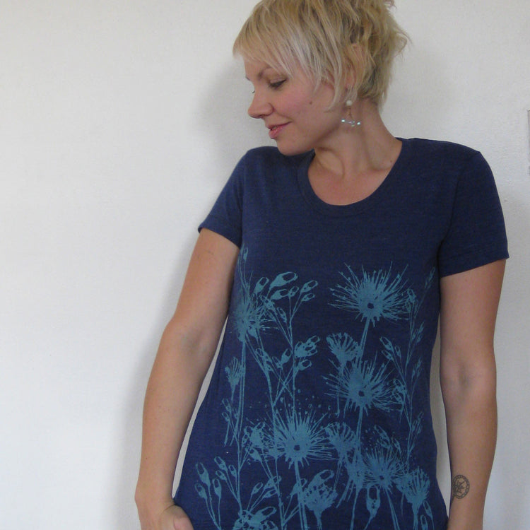A woman wearing a blue t-shirt with a floral design stands against a white wall, looking down at her hand.