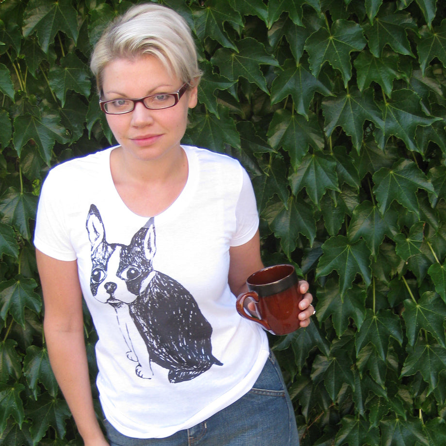 A woman wearing a white t-shirt with a black and white dog graphic is holding a brown mug in front of a green ivy-covered wall.
