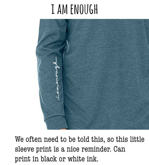 a person wearing a gray long-sleeved shirt with a white text logo on the sleeve. The text reads "I AM ENOUGH" and "We often need to be told this, so this little sleeve print is a nice reminder. Can print in black or white ink."