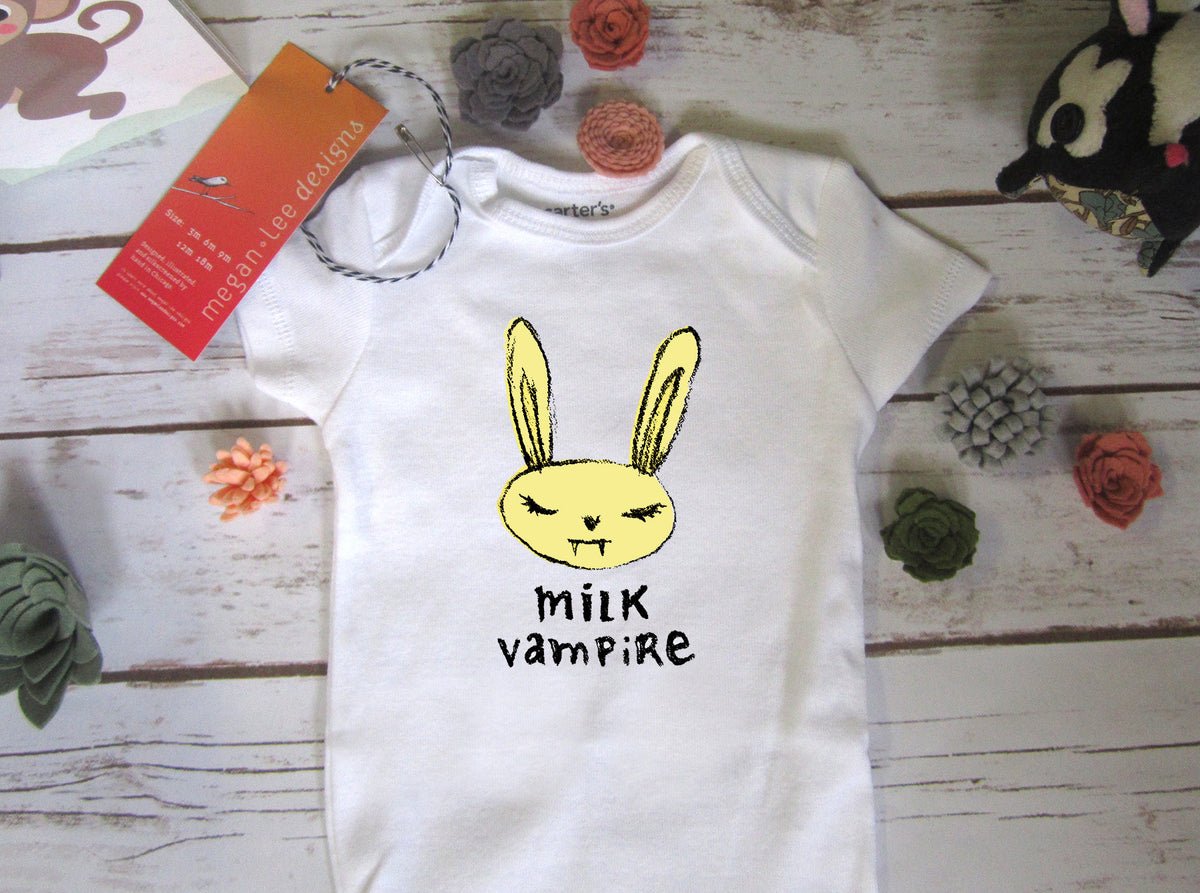 A white baby onesie with a yellow bunny graphic and the text "milk vampire" printed on it, along with a tag and some small decorative items on a wooden surface.